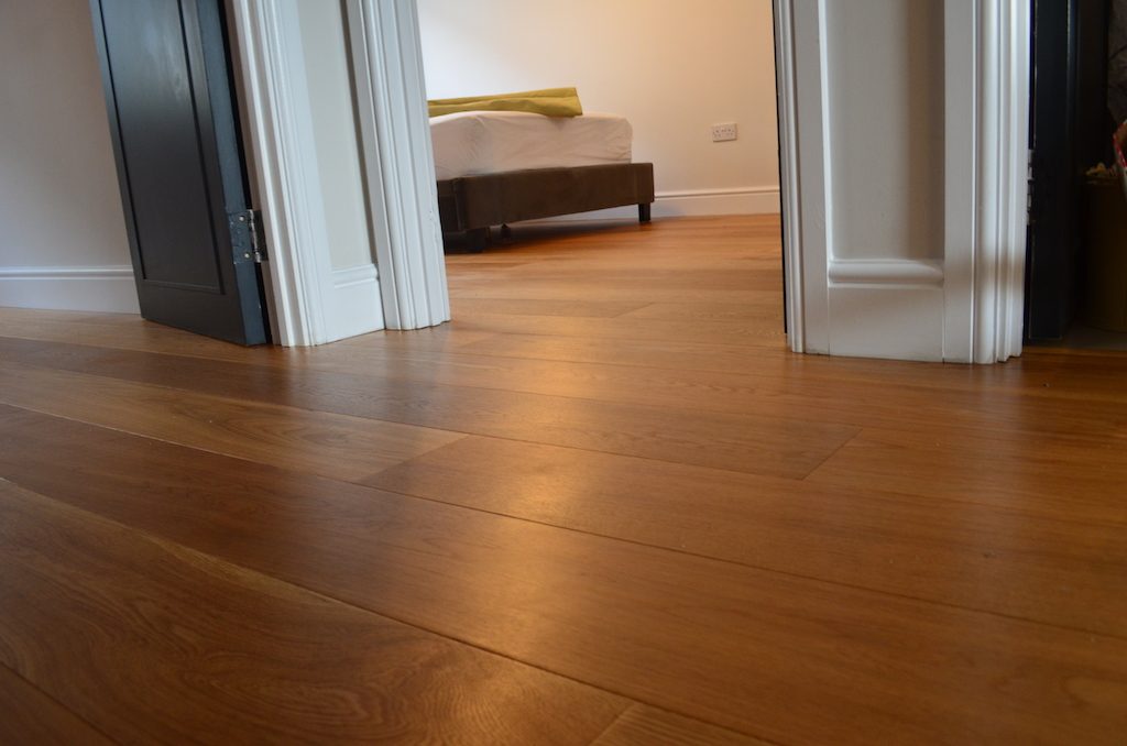 Low view from a landing through a bedroom doorway, honey-toned oak planks running up to the bed