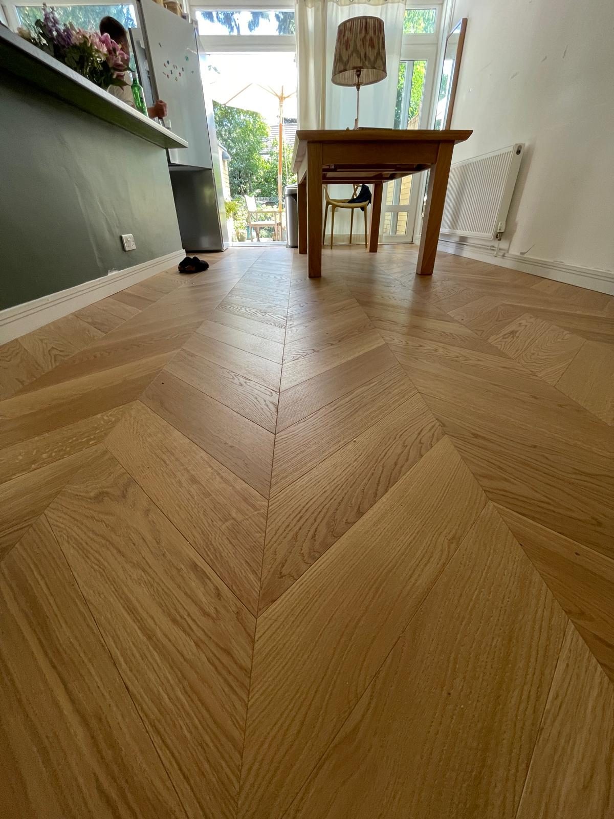 A low view down the centre line of a light oak chevron floor towards a dining table and garden doors